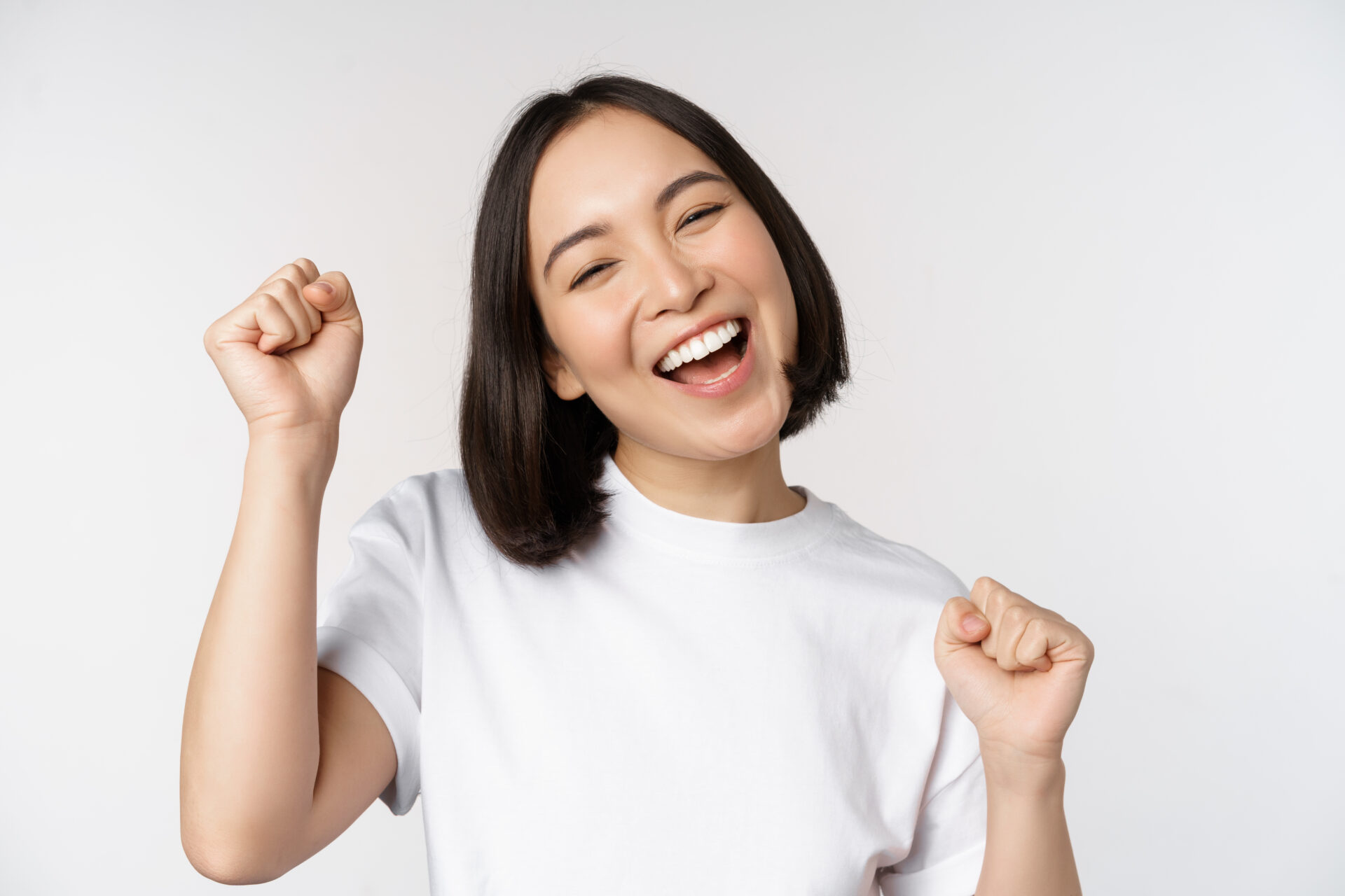 Dancing asian girl celebrating, feeling happy and upbeat, smiling broadly, standing over studio white background